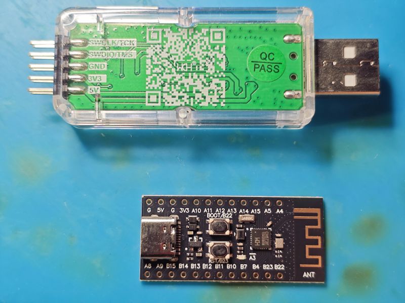 Two electronic modules on a blue surface: a USB programmer with clear casing and a black PCB with microcontroller and antenna.