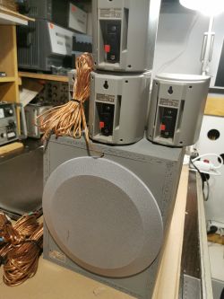 Zestaw szarych głośników z widocznym okablowaniem, ustawiony na półce wśród innego sprzętu elektronicznego.