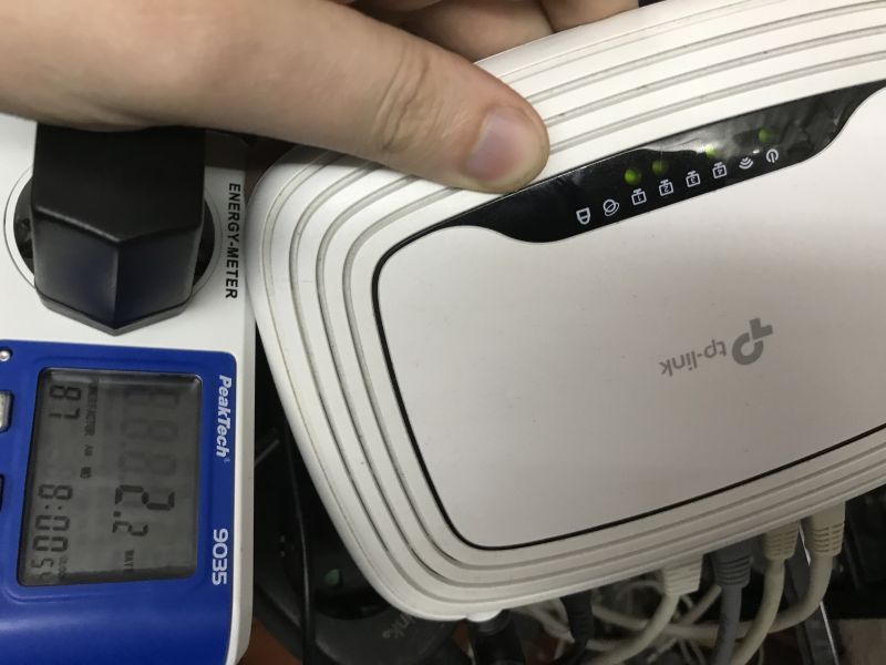 Hand on a TP-Link router connected to an energy meter; green LEDs on, Ethernet cables plugged in at the bottom.