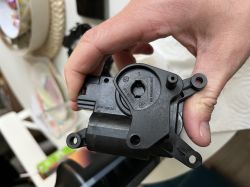 Czarna plastikowa część samochodowa, trzymana w dłoni, na tle stołu z różnymi przedmiotami.