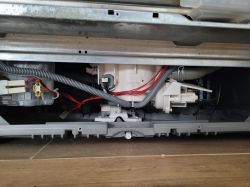 Widok od spodu zdemontowanej zmywarki, widać wiązki przewodów oraz elementy instalacji wodnej.