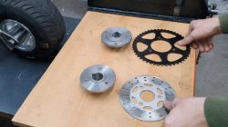 Zwei Hände halten Metallteile neben einem Fahrzeugrad auf einer Werkbank aus Holz.