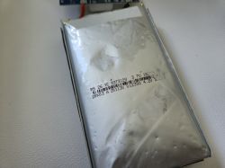 Spuchnięty akumulator litowo-polimerowy na białym tle, z widocznymi oznaczeniami technicznymi.