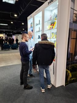Three men talk beside a lit display cabinet with products at a trade show booth.