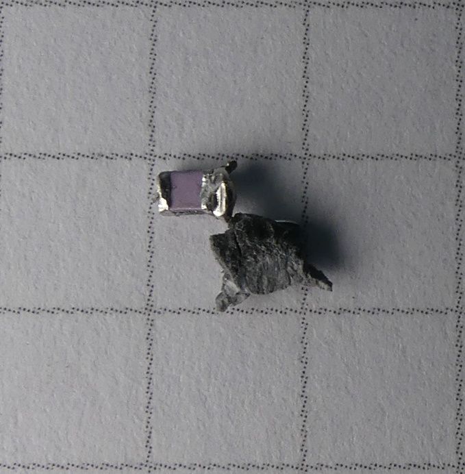 Mały fioletowy kondensator SMD i kawałek szarego metalu na papierze w kratkę