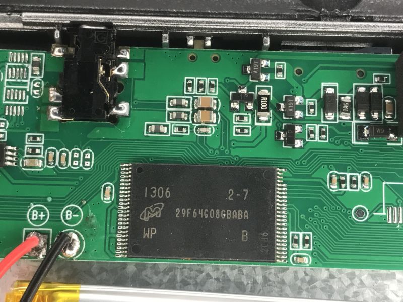 Close-up of a device PCB with chip marked 29F64G08CBABA and solder pads labeled B+ and B-