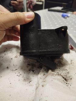 Osoba trzyma czarną plastikową część, prawdopodobnie z urządzenia mechanicznego, na zabrudzonej powierzchni.