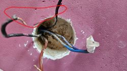 Zbliżenie na puszkę elektryczną w ścianie z widocznymi luźnymi przewodami: dwa czarne, jeden brązowy i dwa niebieskie, z zaznaczeniem czerwonym markerem dwóch czarnych przewodów.