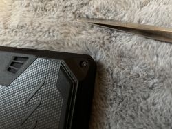 Zbliżenie na uszkodzoną śrubkę w obudowie laptopa oraz metalową pęsetę na szarym futrzanym podłożu.