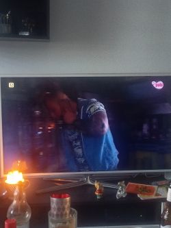 Telewizor Samsung Smart TV z wyświetlanym kanałem TV Puls, na którym widać osobę w koszuli w kwiaty. Przed telewizorem na szafce znajdują się zapalone świece i ozdoby.