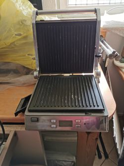 Otwarty elektryczny grill kontaktowy z widocznymi płytami grzewczymi i panelami sterowania, ustawiony na stole.