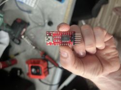 A person holds a red USB to UART converter module with a micro USB port and pin headers.