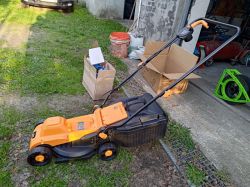 Pomarańczowo-czarna kosiarka elektryczna Ondragon stoi na trawie przed garażem, obok kartonów i narzędzi.