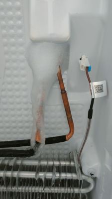 Zbliżenie na wewnętrzną część zamrażarki z widocznym nagromadzeniem lodu na rurce miedzianej, obok czujnika temperatury z niebieską wtyczką.