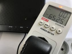 UNI-T power meter with plug, showing 0.0 W and 65.25; black monitor in the background