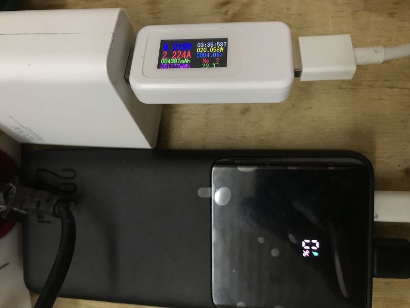 USB power meter with display between a charger and cable, next to a power bank with a small screen on a wooden desk