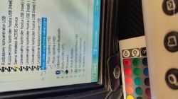 Widok ekranu komputera pokazujący menedżer urządzeń, na którym widać różne urządzenia USB. Obok leży pilot z kolorowymi przyciskami.