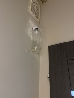 Plastikowa butelka podłączona do metalowej rurki przy kratce wentylacyjnej na ścianie.