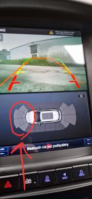 Ekran nawigacji samochodowej w Fordzie Focus MK3 z widokiem z kamery cofania i wskazaniem czujników parkowania.