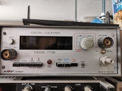 Licznik cyfrowy AMF Venner model 7736 stojący na półce wśród sprzętu elektronicznego.