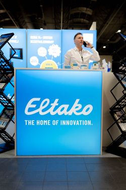 Eltako trade show booth with a blue illuminated panel and a person on the phone behind the counter.