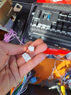 Osoba trzyma w ręku małe białe złącza do radia samochodowego, w tle widoczny jest wzmacniacz.