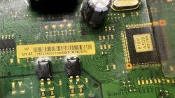 Zbliżenie na płytę elektroniczną z widocznym chipem, kondensatorem i żółtą etykietą z kodem kreskowym.