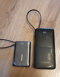 Dwa czarne powerbanki Baseus na drewnianym stole, wyświetlacze 100% i 82%, podłączone kable USB
