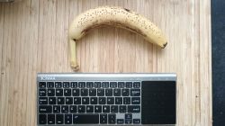 Mała klawiatura bezprzewodowa z touchpadem obok banana na drewnianym blacie.