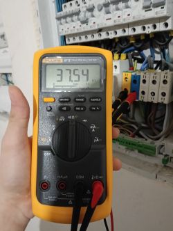 Multimetr pokazujący pomiar rezystancji 37,54 kΩ na tle skrzynki elektrycznej.