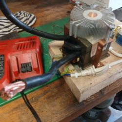 Transformator z aluminiowym radiatorem i grubymi przewodami na blacie obok czerwonej ładowarki