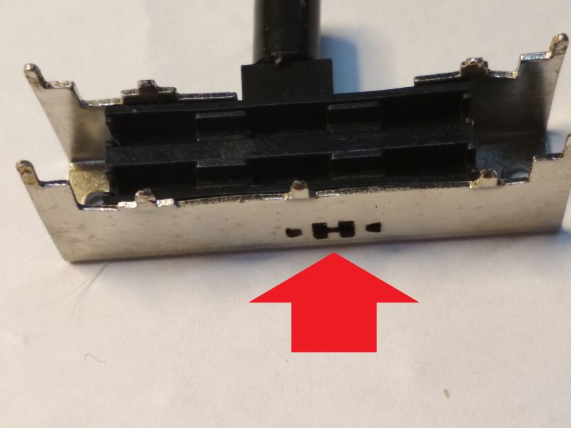 Zbliżenie na rozebrany metalowy przełącznik suwakowy z widoczną czarną częścią plastikową i czerwoną strzałką wskazującą na otwór w obudowie.