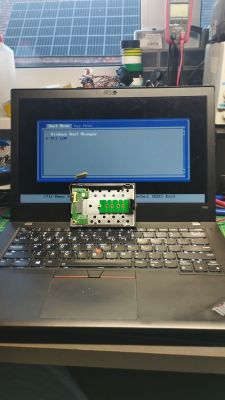 Laptop z otwartym boot menu i wyjętym adapterem dysku SSD leżącym na klawiaturze.