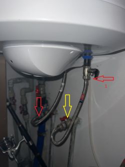 Zbliżenie na instalację hydrauliczną z metalowymi wężykami, oznaczonymi strzałkami: żółta strzałka wskazuje na miejsce podłączenia.