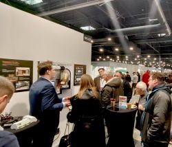 Trade show booth in a hall; people talk around tall tables near wall displays and a mounted device.