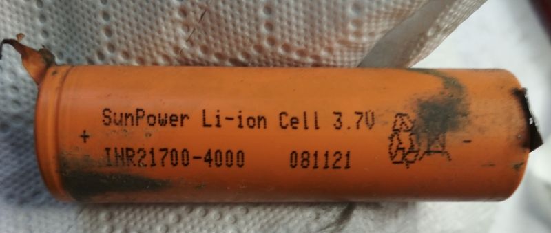 Pomarańczowe ogniwo litowo-jonowe SunPower INR21700-4000 z widocznymi śladami zabrudzeń i uszkodzeń izolacji, leżące na białej tkaninie.