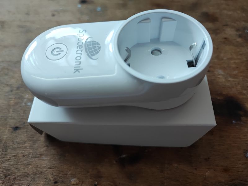 Biała inteligentna wtyczka Spacetronik leżąca na białym pudełku, na drewnianej powierzchni.