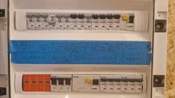 Widok rozdzielnicy elektrycznej z wyłącznikami i opisami na niebieskiej taśmie