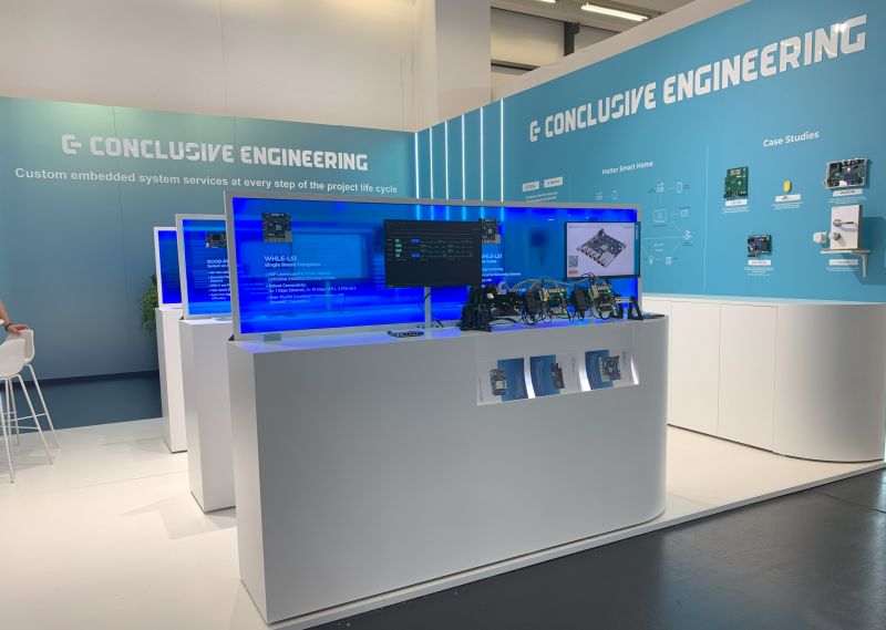 Stoisko Conclusive Engineering z różnymi urządzeniami i ekranem prezentującym usługi.