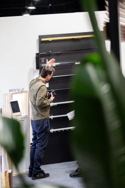 Man at a trade show booth examines a large black wall panel; blurred green leaves partly block the view.
