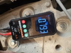 Multimetr cyfrowy z wyświetlaczem 27,93 V DC leżący na metalowej powierzchni z śrubami