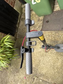 Kierownica hulajnogi elektrycznej Xiaomi 3 Lite na tle betonowej nawierzchni i zielonych pojemników.