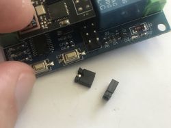Close-up of a PCB with a relay and buttons; two small black jumper caps lie beside it.