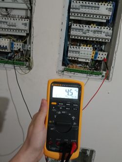 Ręka trzymająca multimetr pokazujący napięcie 4,5 V na tle dwóch rozdzielnic elektrycznych.