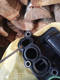 Uszkodzony plastikowy kolektor dolotowy chłodzony płynem, z narzędziem wskazującym na jego pęknięcie, na tle drewna opałowego.