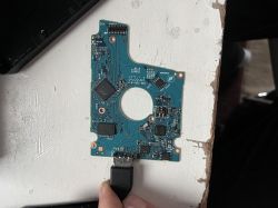 Płytka PCB z dysku twardego Toshiba z podłączonym kablem USB, leżąca na białym stole.