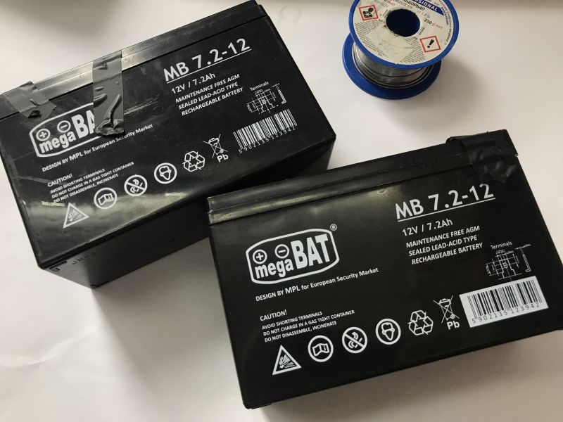 Two black 12V 7.2Ah gel batteries from MegaBAT and a spool of soldering tin on a white background.