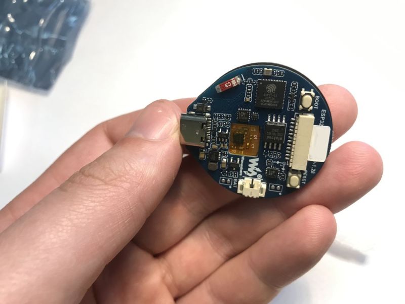 Okrągły moduł elektroniczny ESP32-S3-Touch-LCD-1.28 trzymany w palcach, widok od strony złączy i elementów elektronicznych.