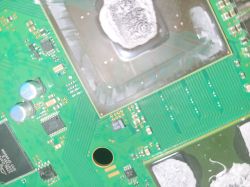 Zbliżenie na płytę główną konsoli PS3 Slim J001 z widocznym procesorem CELL po delidzie oraz potencjalnymi uszkodzeniami ścieżek.