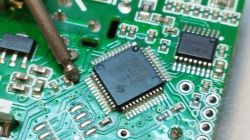 Microcontroller being soldered onto a green circuit board with visible traces and components.
